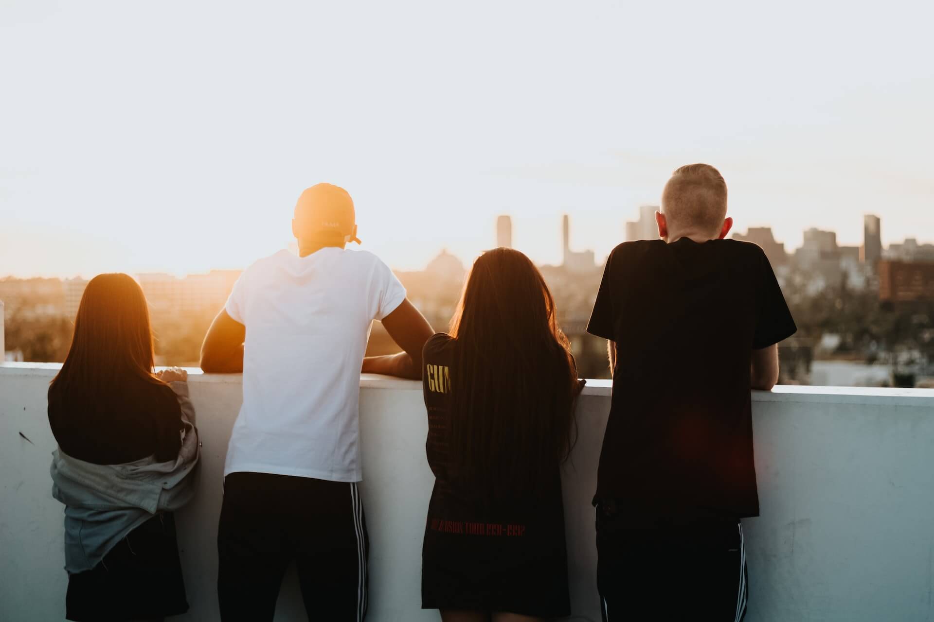four young persons shown fromteh back looking out over a short wall observicing a city skyline at sunset