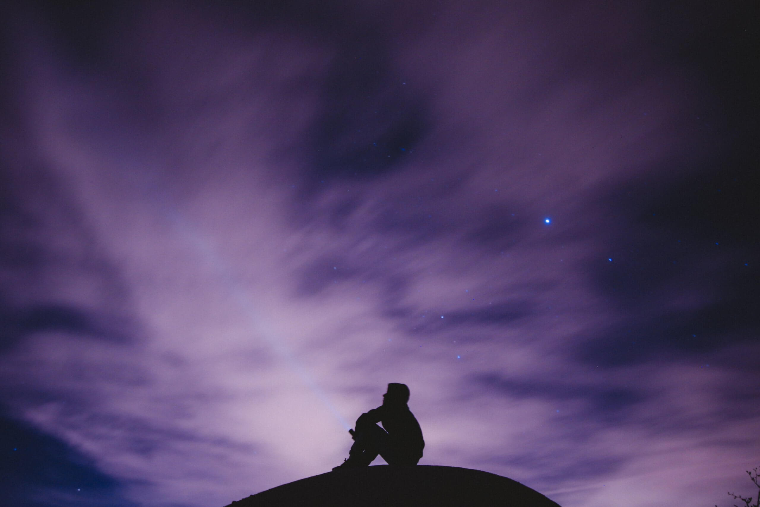 Silhouette of youth siting on rock pointing flashlight up into dark sky with purple clouds and a few stars and plants