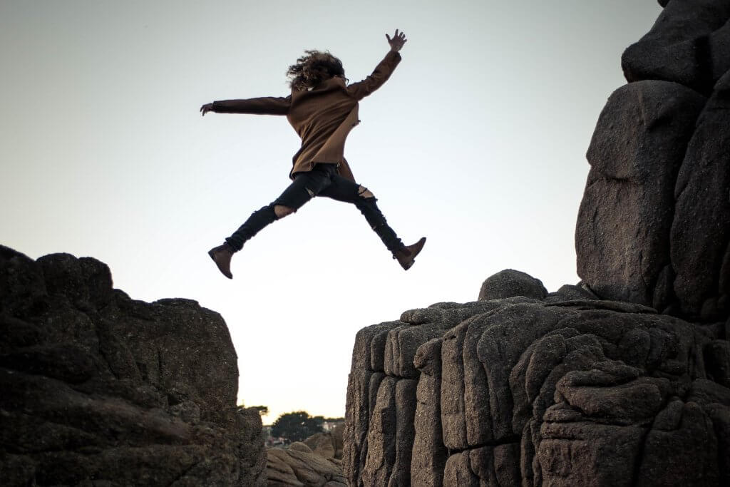 person silhouetted against sky leaping from rock formation to another rock formation - photo