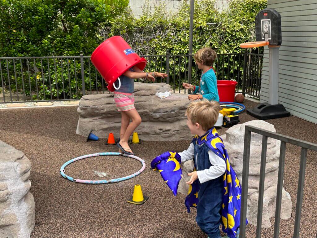 children playing in UUSM play yard; one child wears wizard robe and another has a giant red bucket on her head - photo