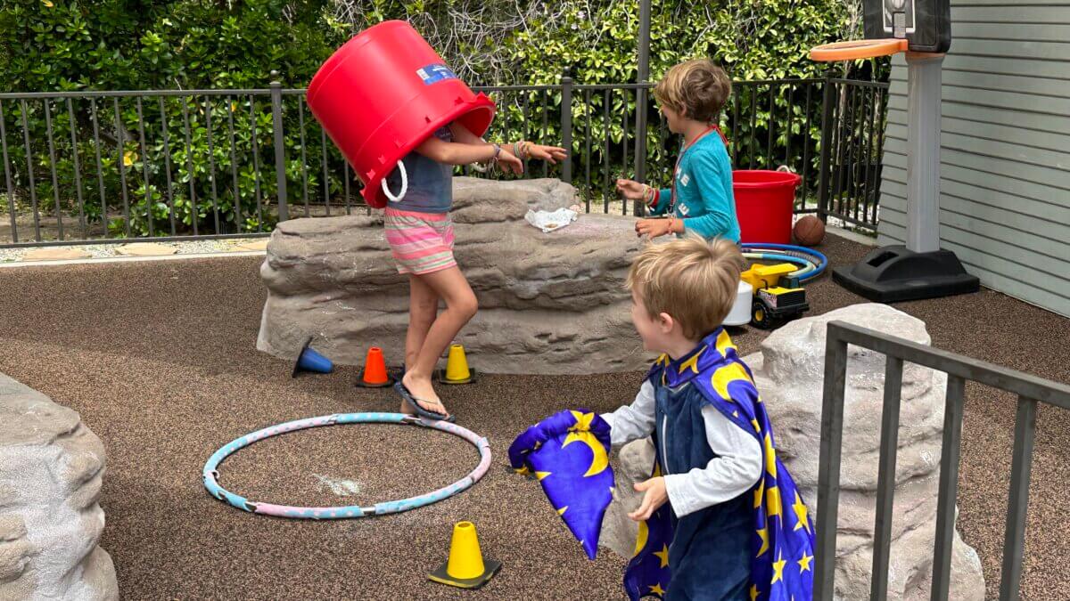 chilren playing in UUSM play yard; one child wears wizard rbed and another has a giant red bucket on her head - photo